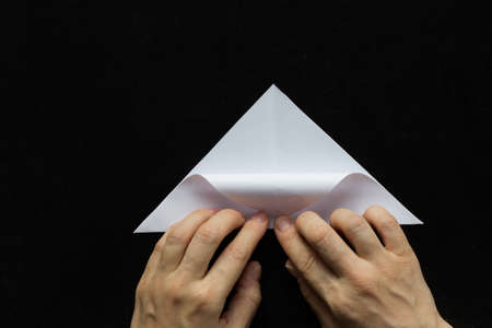 Making snowflake. Step 4. Woman hands folding corner of paper triangle to bottom to mark line on black backgroundの写真素材