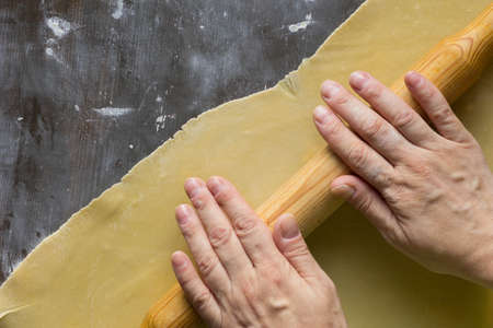 Woman hands rolling dough for noodles by rolling pin on the powdered by flour wooden surface