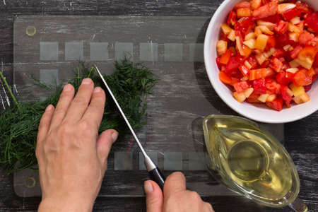 Hand of woman cutting fresh dill with knife for salad on glass board on black backgroundの写真素材