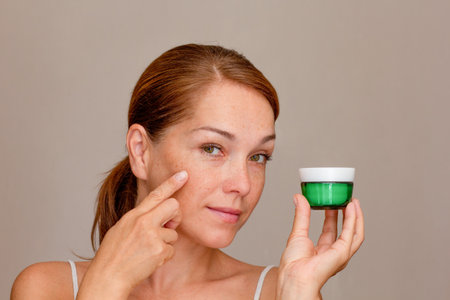 Portrait of middle aged woman with freckles showing on skin by index finger and holding creamの写真素材