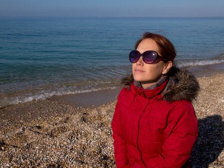 Caucasian woman in red puffer coat on sea beach looking up at sun and sky in sunglassesの写真素材