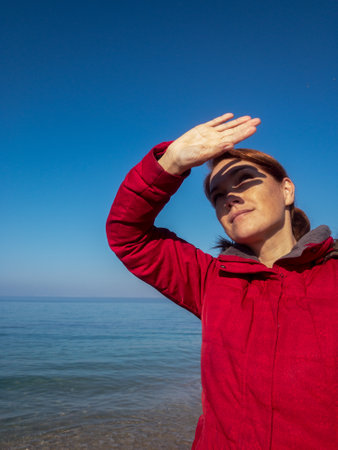 Caucasian woman in red puffer coat on sea beach looking up close her face by hand hiding from sunlightの写真素材