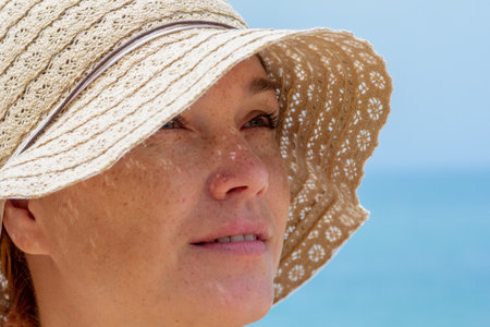 Close up caucasian middle aged woman face in sun hat looking aside over blue seaの写真素材
