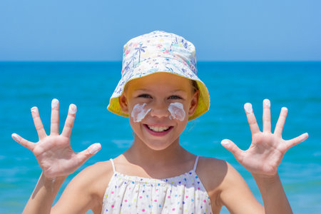 Happy girl at the beach, wearing a colorful sun hat, with sunscreen applied on her face and hands...