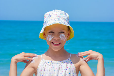Child happily applying sunscreen on her face and shoulders at the beach, wearing a vibrant hat, with the ocean waves and bright sky creating a joyful summer atmosphereの写真素材