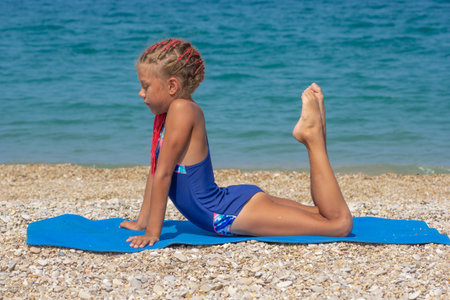 A young child does Swan Pose on a sandy beach, pretending to be a seashell in a fun and imaginative...