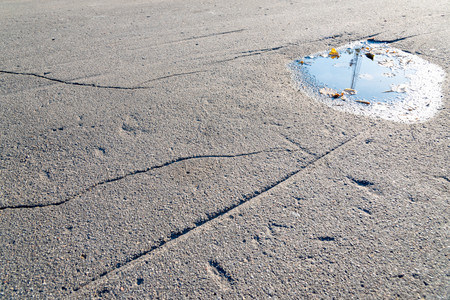 A puddle of water on the road surface (asphalt) with dry leaves on the edges. Blue sky and sun are...