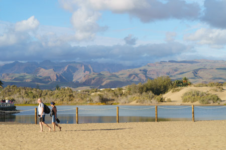 Maspalomas, Gran Canaria, Spain - November 21 2018: Sandy beach of Maspalomas. Heterosexual couple walk along. Sunny summer day. Tourists' concept, side view. beautiful volcanic landscapeのeditorial素材