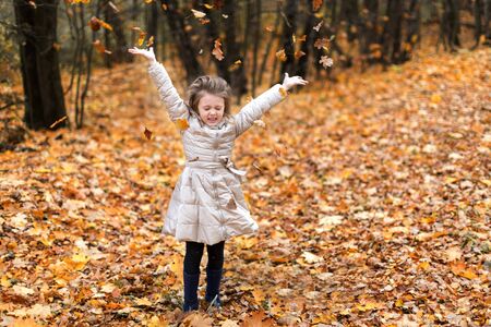 A pretty blonde little girl in the forest throws autumn leaves. Fun in the fresh air in autumn. The teenager smiles happily.の写真素材