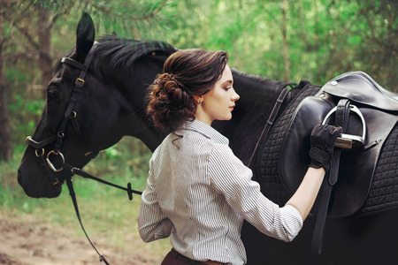 Beautiful girl rider with a black horse, dressed in a light shirt in a green forest Park. The concept of horse riding.の写真素材