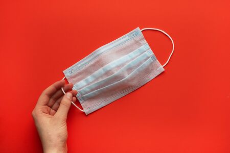 Medical mask in female hands on a red flat background lay top view. Protection against viral...