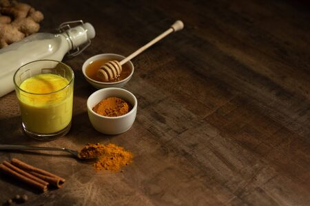 Turmeric Milk is poured into a glass. Next to it are the ingredients for making a drink - milk in a bottle on a napkin, spices and honey. Golden drink on dark moody background, selective focus. Top viewの写真素材
