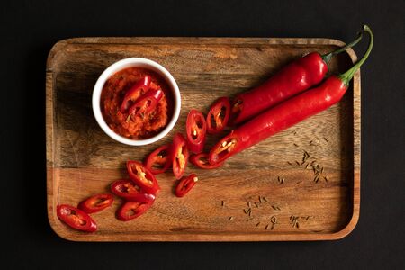 Harissa sauce in the gravy boat. Ingredients for cooking - chili peppers, garlic and spices. Everything lies on a wooden tray. Black background and top view.の写真素材