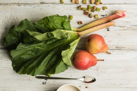Rhubarb stalks and two pears on a white wooden background. A mug of sugar and a metal spoon lie nearby. Complementary colors.の写真素材