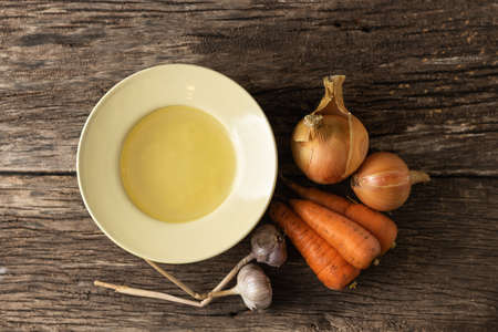 Bowl with clear rich broth. White crockery on an aged wooden table. Vegetables - carrots, onions, garlic, spicesの写真素材