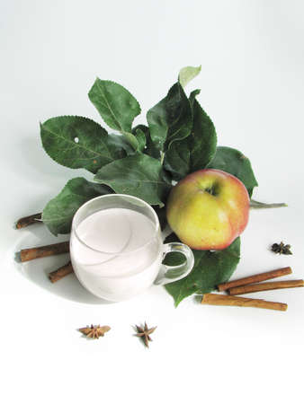 Clear glass cup with cream, ripe apple on an apple tree branch, cinnamon, and star anise on white tableの写真素材