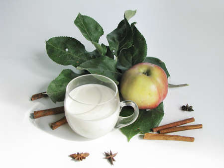Clear glass cup with cream, ripe apple on an apple tree branch, cinnamon, and star anise on white tableの写真素材