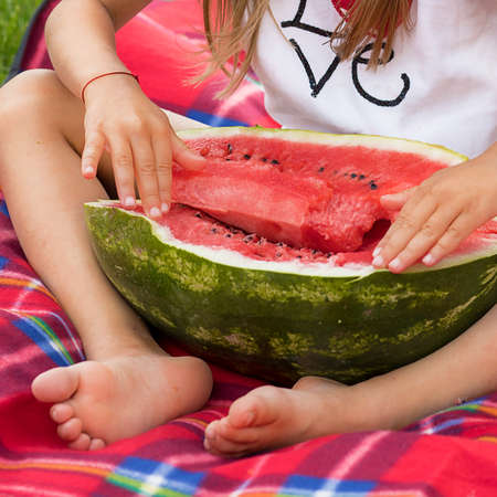 Photo of a half of a ripe huge watermelon, which is hugged by the hands and feet of a little girl. Juicy ripe fruit. Summertime. Harvest. Kids Vacation and picnic on the grassの写真素材