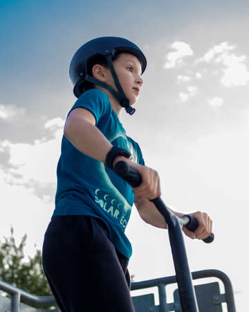 Boy 10 years with helmet on head stands on ramp of skatepark against gloomy sky holding scooter With...