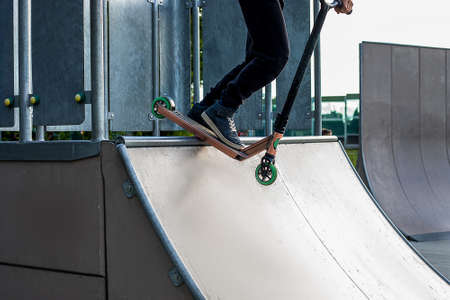 Teenager boy rides on kick scooter in outdoor skatepark. Summer extreme sports outside. Feet of male...