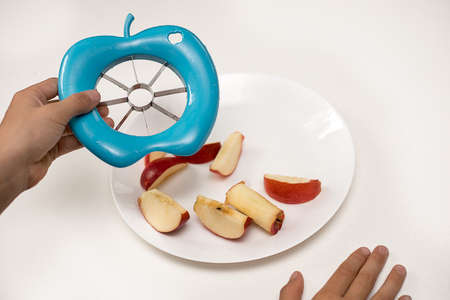 Red Apple cut into pieces with special cutting tool device Easy Corer or Slicer in blue color in kids hands. On white background table on plate. Close up photoの写真素材