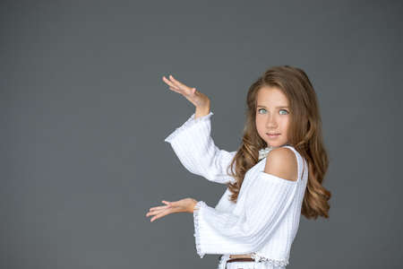 A beautiful baby girl points to an empty space on gray background for your text or subject. Fashionable casual light clothing, natural look. Presentation and education concept. Studio shotの写真素材