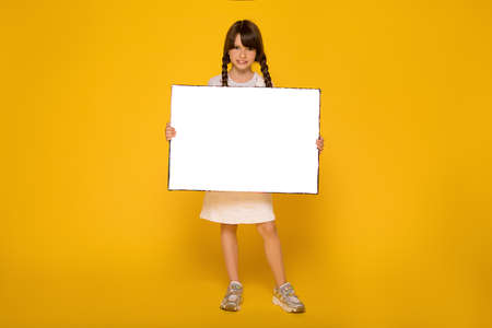 Beautiful School girl holding white sheet. Cute kid with paper. Yellow background. Copy spase. Child holding empty board for text advertisingの写真素材