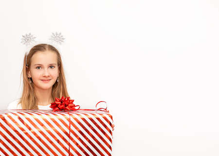 A beautiful kid girl holding a Christmas red striped gift box with a ribbon and a bow in her hands. There are snowflakes on the head. Isolated on white. Studio phpto. Copy spaceの写真素材