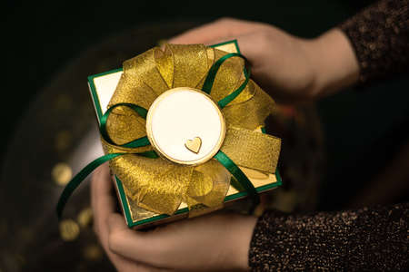 Female hands in swetear holding gift box wrapped in green paper with gold bow. Congratulations on a holiday for a man. Mock up. Copy space.の写真素材