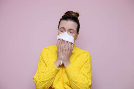 Photo ill woman in a sweater sneezes into a napkin on a pink background, allergic rhinitis. The concept of colds, seasonal illnesses, flu.の写真素材