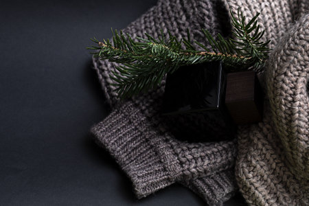Male perfume on a black background in a black glass jar with a wooden lid. Nearby is a coarse-knit sweater and coniferous branch. Gift conceptの写真素材