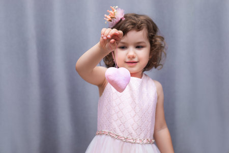 A pink soft toy in the shape of heart in the hands of a 4-5 year old girl. Valentine's Day for Valentine's Day. Close-up portrait on gray backgroundの写真素材
