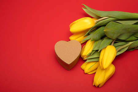 Concept of congratulations on holiday Moms Day, Valentines Day. Yellow tulips and heart-shaped gift box made of craft cardboard on red background. Copy space, mock upの写真素材