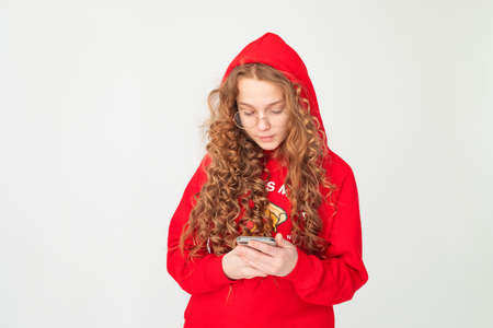 Red haired curly hair teenage girl in red hoodie with hood, using reading smartphone on white background. Concept of online communication, education. Looks at phoneの写真素材