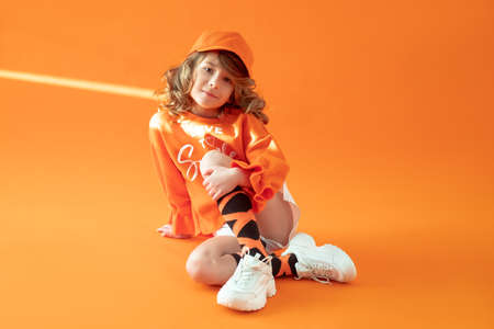 Girl 6 years old, sitting on the floor in studio on orange background, posing in casual clothes, cap and sneakers. Childhood, fashion, advertising, sports. Close up, copy space. Sunbeam on headの写真素材