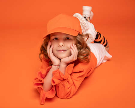 Smiling little girl in cap and fashionable clothes lying down on floor in studio on orange background. Mock upの写真素材