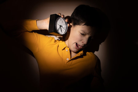 teenage child boy holding alarm clock in hands and fall asleep on the go, night. Concept of sleep disturbance in children and adolescents. Hard light and close-up photoの写真素材