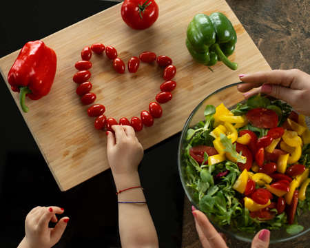 Children's hands lay out heart of cherry tomatoes on wooden board among vegetables. Cooking process with child. Top viewの写真素材