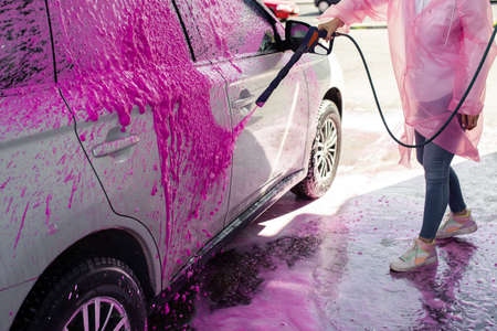 Unrecognizable woman in pink raincoat washes car at self-service car wash, stock photo