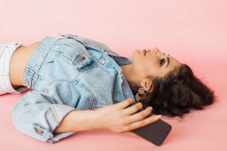 Beautiful young woman lies on pink background in studio with phone in her hands. loneliness and disappointment concept.の写真素材