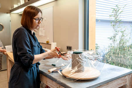 Brunette woman in glasses in casual clothes cuts polythene packaging with scissors. Bouquet in box...