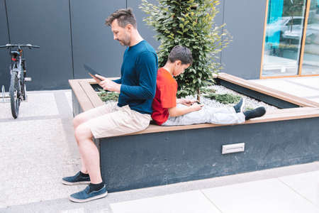 Father and son spend time in the city park, sitting back to back and using a smartphone or tablet. Modern lifestyleの写真素材