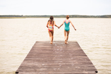 2 girls on holiday running on wooden bridge to jump into water, rear view, summer time, vacations, holding handsの写真素材
