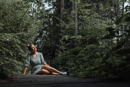 Woman teacher sits on wooden bridge in park and enjoys singing of birds and contemplates nature. Concept of pleasure and relaxation in silence.の写真素材