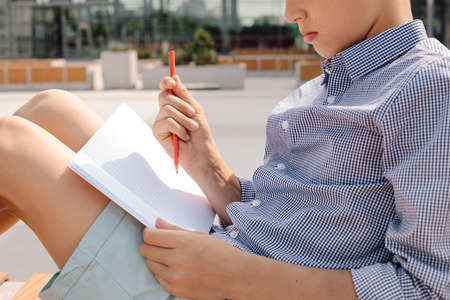 Photo of a boy working in notebook with pencil on bench in schoolyard. Informal educational...