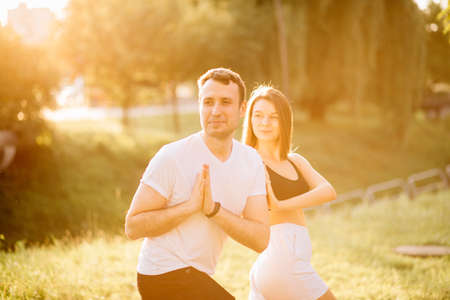 Young couple man and woman doing sports, yoga on city lawn, summer evening, stretching together on sunset, concentrationの写真素材