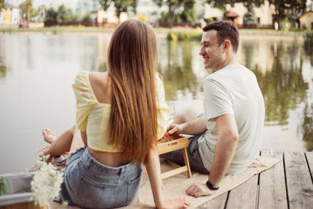 Rear view of young couple on picnic near river or lake, woman and man drinking wine outdoors together, people having fun on vacationの写真素材