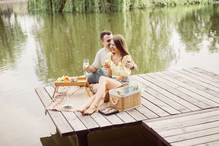 Side view of young happy couple on romantic picnic near river or lake, woman and man drinking wine outdoors together, people having fun on vacationの写真素材