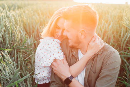 Happy young couple embraces, sitting in tall grass in field in rays of setting summer sun. Love and tenderness. Beautiful moments of life. Youth and beauty. Peace and carelessness.の写真素材