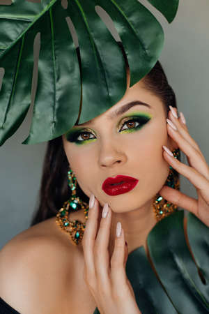 Close up face of beautiful young woman covering by green monstera green palm. Portrait of beauty woman with makeup red lips clear skin with big green glossy leaves.の写真素材